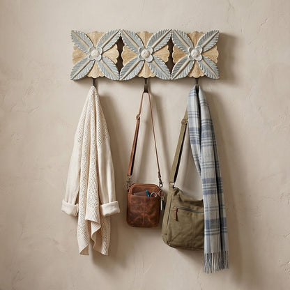 A decorative woven wall hook rack with three hanging hooks, styled in a warm, boho-chic entryway. The rack features a cream and light blue geometric pattern with three floral motifs.